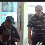 A still from surveillance video shows two men, one wearing a baseball cap with yankees logo and the other wearing glasses and a short-sleeved polo shirt that is dark with light white horizontal stripes