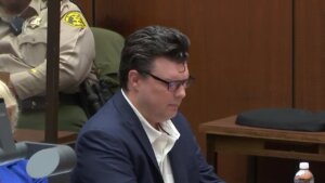 A man wearing a suit without a tie sits in a courtroom looking down