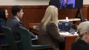 A man sits next to a woman in court with a computer on the table and a screen in the background