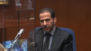 A man in suit and tie sits behind plexiglass and a microphone