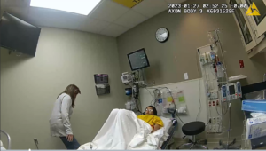 a still from police bodyworn camera shows a woman laying a hospital bed and another woman standing by a wall