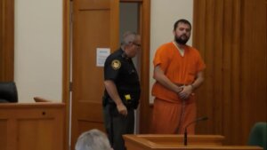 A man in orange jail uniform enters court in shackles