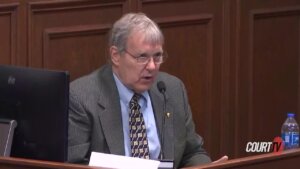 A man in a suit sits with papers in front of a microphone as he's testifying