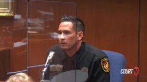 A man in a fire dept uniform speaks into the microphone from the witness stand