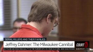 A man in glasses and an orange prison jumpsuit stands in court