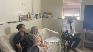 A man sits in a chair with a laptop on his lap in front of microphones in a living room