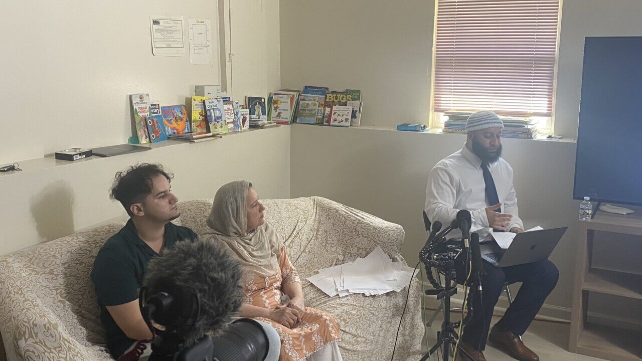 A man sits in a chair with a laptop on his lap in front of microphones in a living room