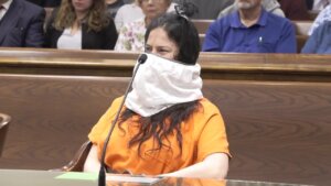 A woman in an orange prison jumpsuit sits with a white cloth over her face in court