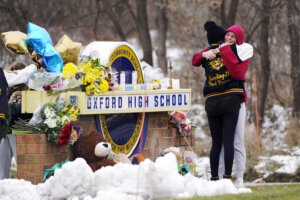 A brick sign that says 'Oxford High School' is adorned with flowers and candles and two people hug in front of it. There is snow on the ground.