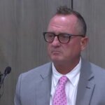 A man in a gray suit with a pink tie testifies in court