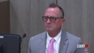 A man in a gray suit with a pink tie testifies in court