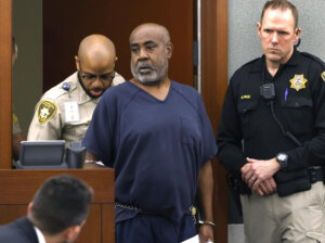 A man in a blue shirt stands in court, flanked by deputies