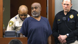 A man in a blue shirt stands in court, flanked by deputies