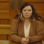 Curly-haired woman in a brown cardigan and white tee shirt sits in court.