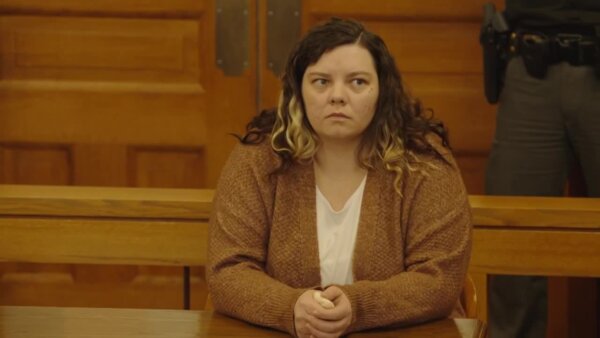 Curly-haired woman in a brown cardigan and white tee shirt sits in court.