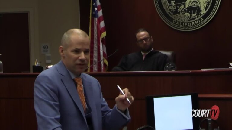 A bald, middle-aged attorney wearing a purple suit and orange tie argues in court.