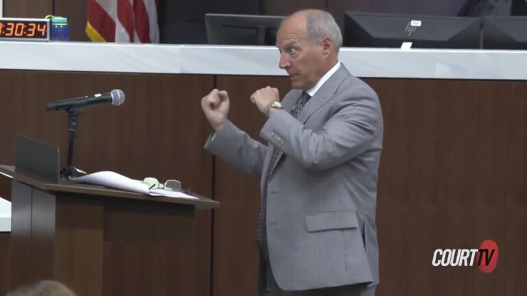 A balding male attorney in a grey suit stands at a podium.