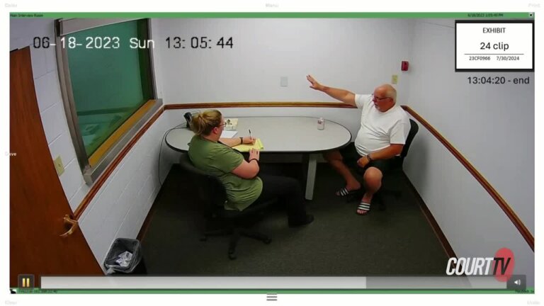 Large older man in a white tee shirt sits in an interrogation room.