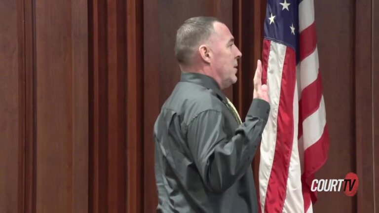 witness swears in while holding up his right hand while standing at the witness box next to an American flag