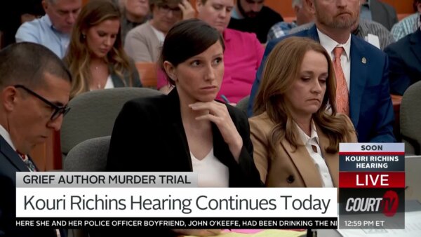 Dark-haired young woman sits in court wearing a black blazer and white scoop neck blouse.