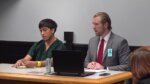 Man dark hair sits next to his attorney with blonde hair and red tie