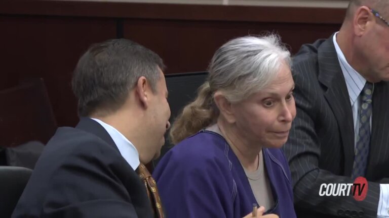 Elderly woman at defense table wears a purple prison jumper.