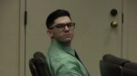 Young male defendant sits in court wearing glasses and a green button down dress shirt.