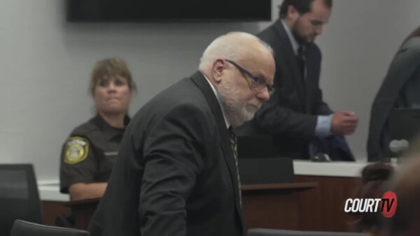 Defendant at defendant's table in court glances at the camera. He's wearing a suit.