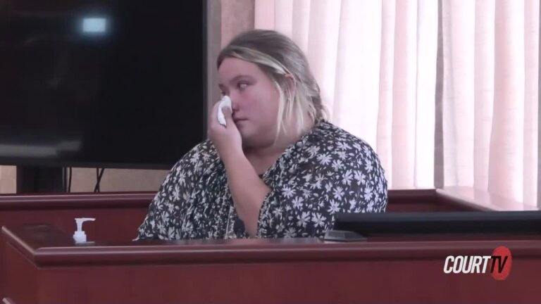 Young, blonde female witness in a floral print blouse clutches tissues on the stand