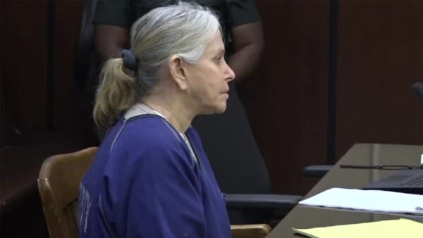 Elderly female defendant in a purple prison jumper sits at defense table.