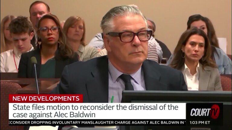 Older gentleman with grey, slicked-back hair, sharp suit and thick black glasses sits at defense table