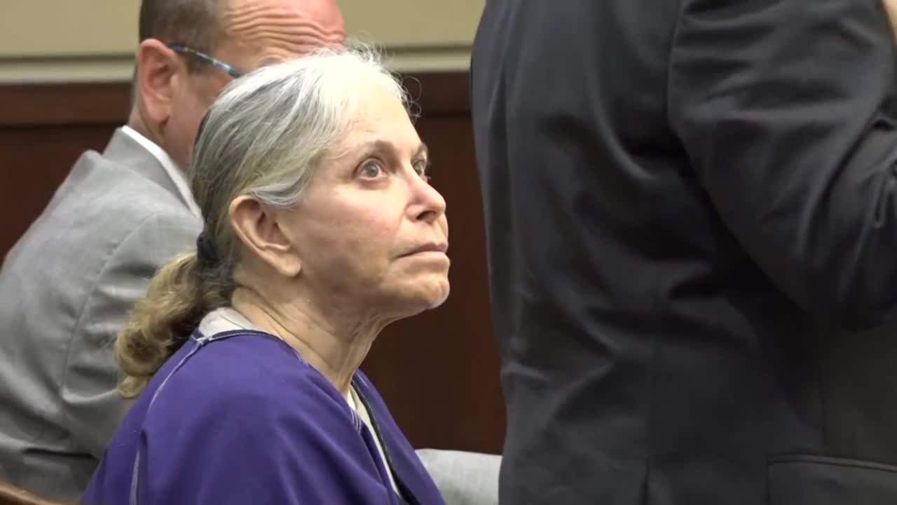 Elderly woman in prison jumper sits at defense table.