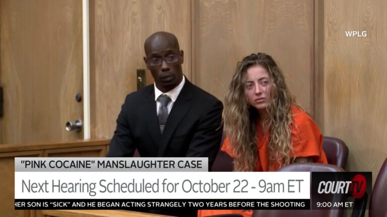 Young blonde curly-haired defendant wears an orange prison jumpsuit as she sits next to her lawyer.