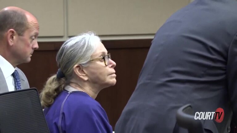 Elderly woman in glasses and prison garb listens in court.