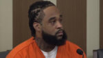 Bearded young man in orange prison jumper sits in court.