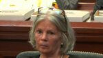 Older female defendant wears pearls and a black dress. Her grey hair is styles in a bob.