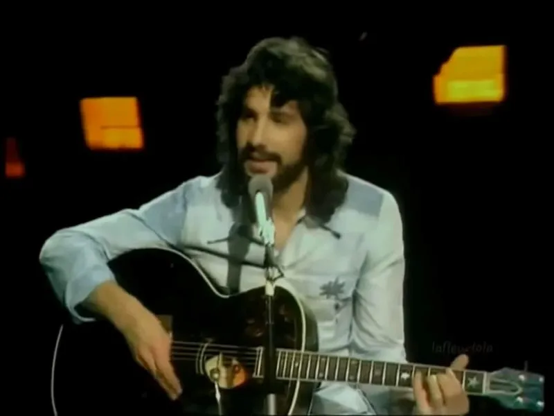 Cat Stevens seated with an acoustic guitar against a dark backdrop speaks into a microphone.
