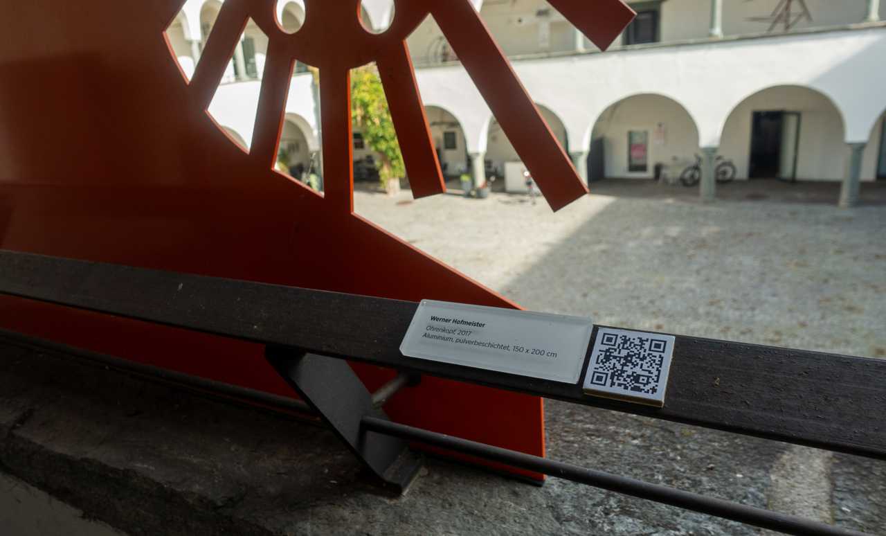 An information label for the sculpture Ohne Titel (2017) by Werner Hofmeister, detailing its material and dimensions, displayed in a sunlit courtyard.