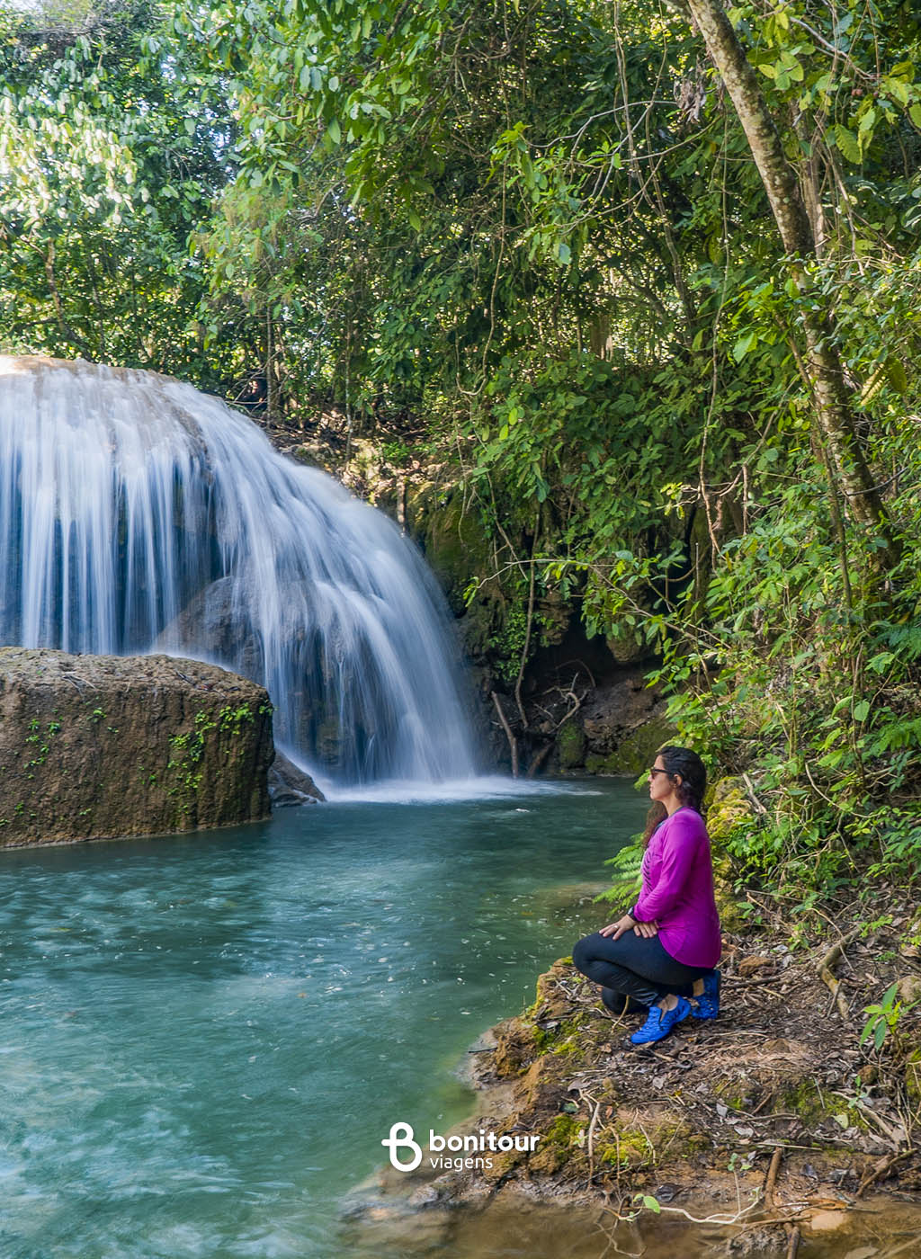 Mulher sentada em beira de rio contempla uma cachoeira na Estância Mimosa.