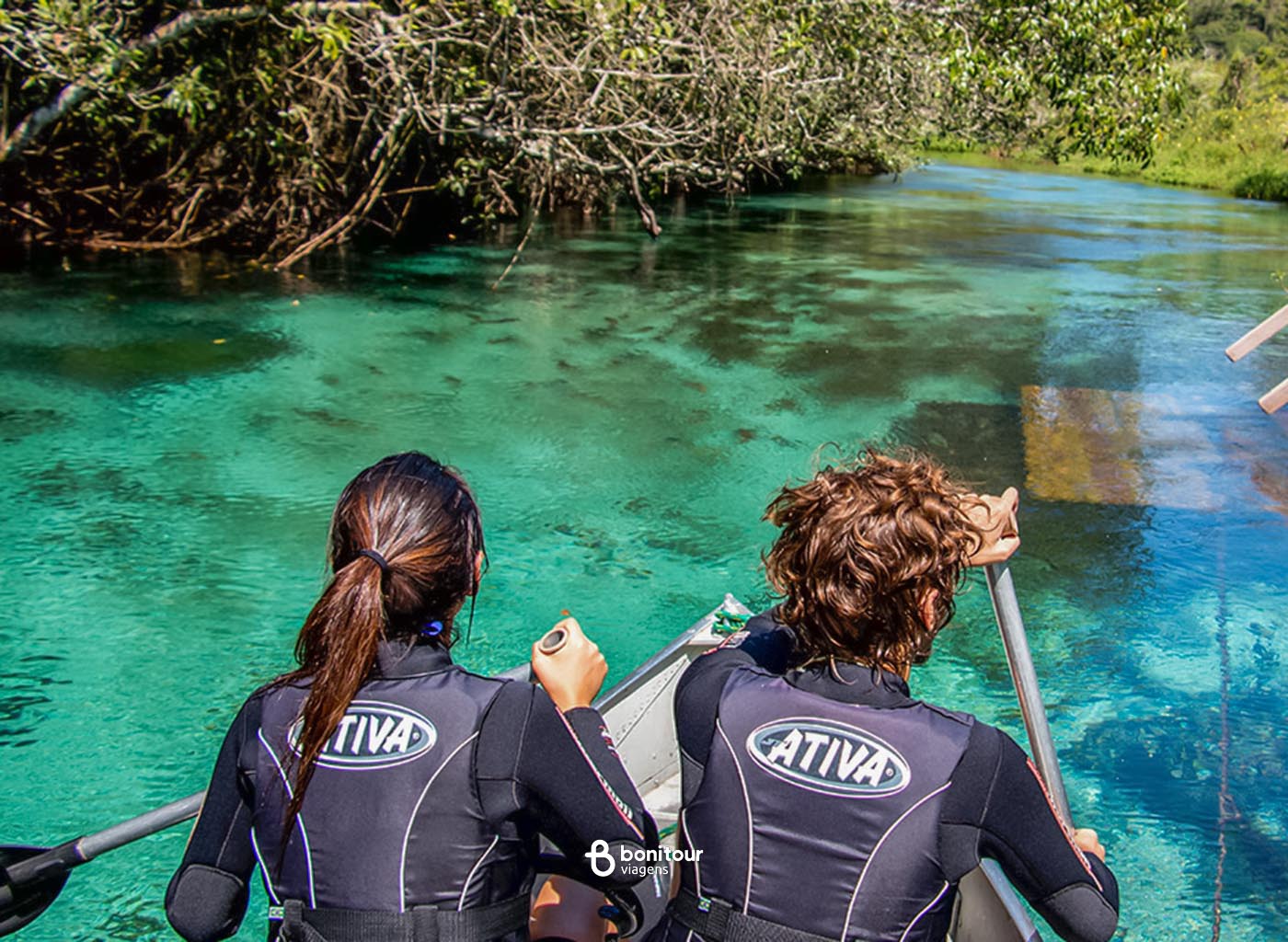 Duas pessoas  de costas para a foto remando em barco pelo Rio Sucuri, com vista para as águas cristalinas do rio e densa vegetação nas margens.