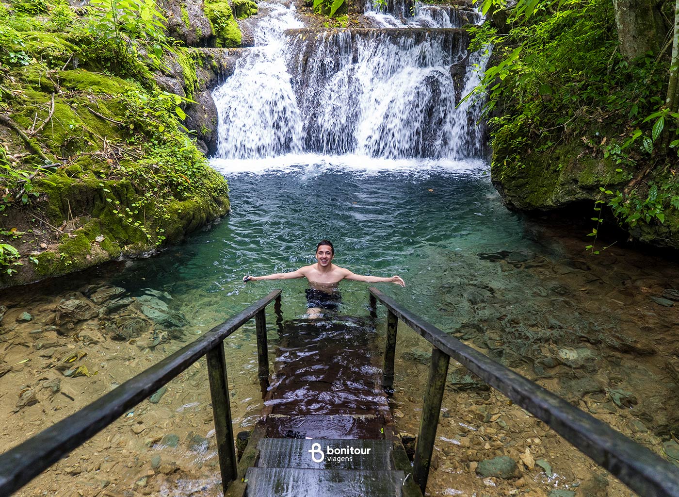 Pessoa em deck de madeira parcialmente submerso, com cachoeiras da Boca da Onça