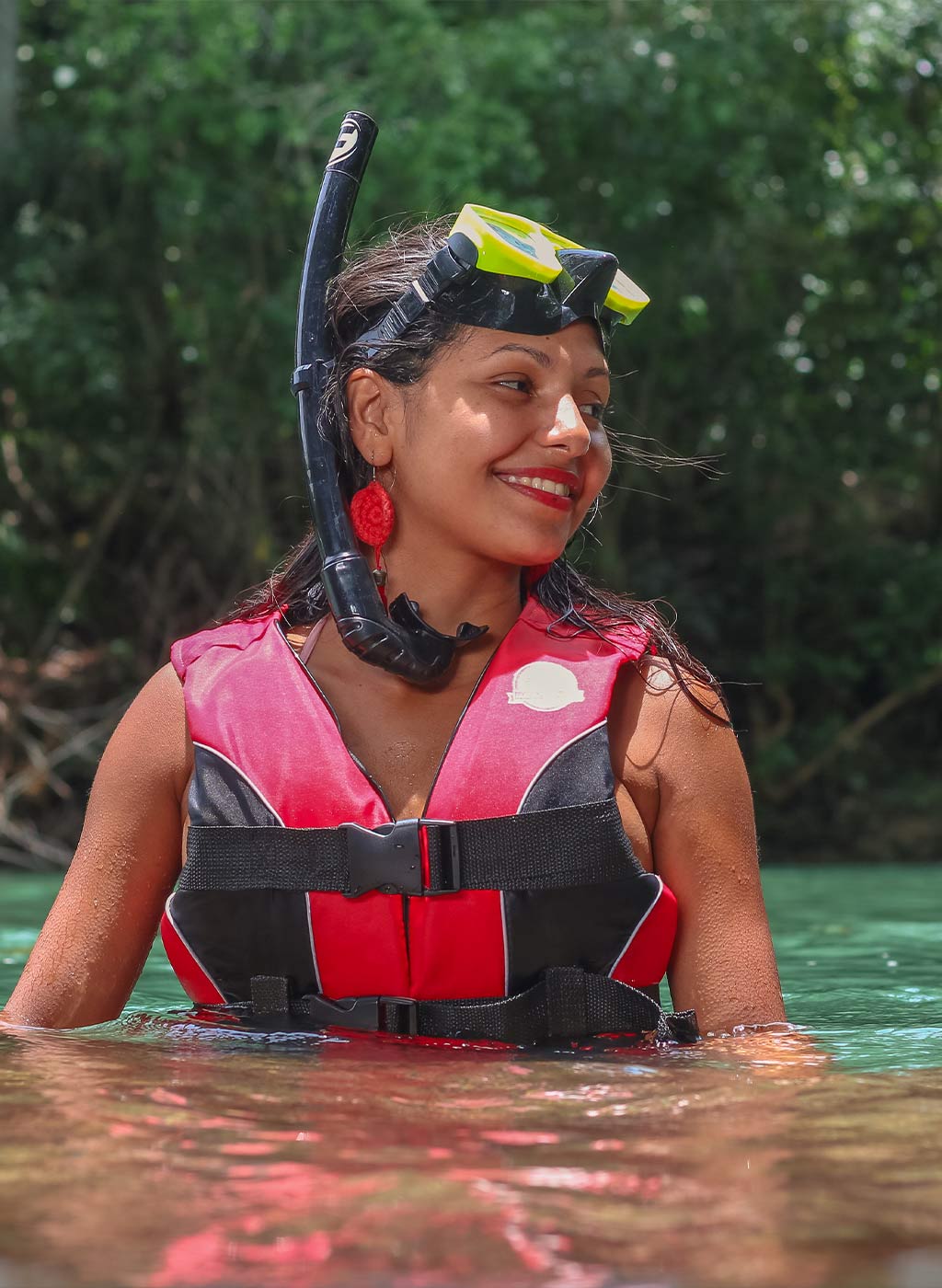 Pessoa com colete salva vidas e óculos de mergulho acima dos olhos, sorrindo, com água até a cintura e vegetação ao fundo