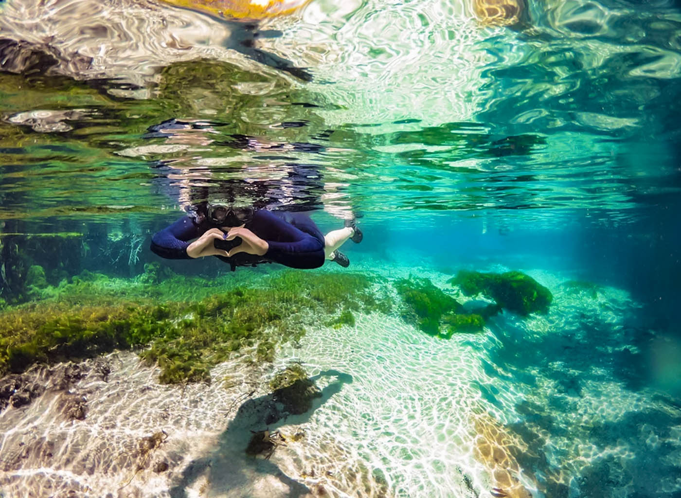 Pessoa com equipamento de flutuação, dentro das águas cristalinas do Rio Sucuri, fazendo coração com as mãos;