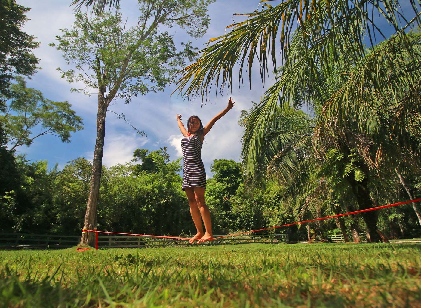 Mulher se equilibrando em cima de slackline  com os braços para cima, com natureza ao fundo