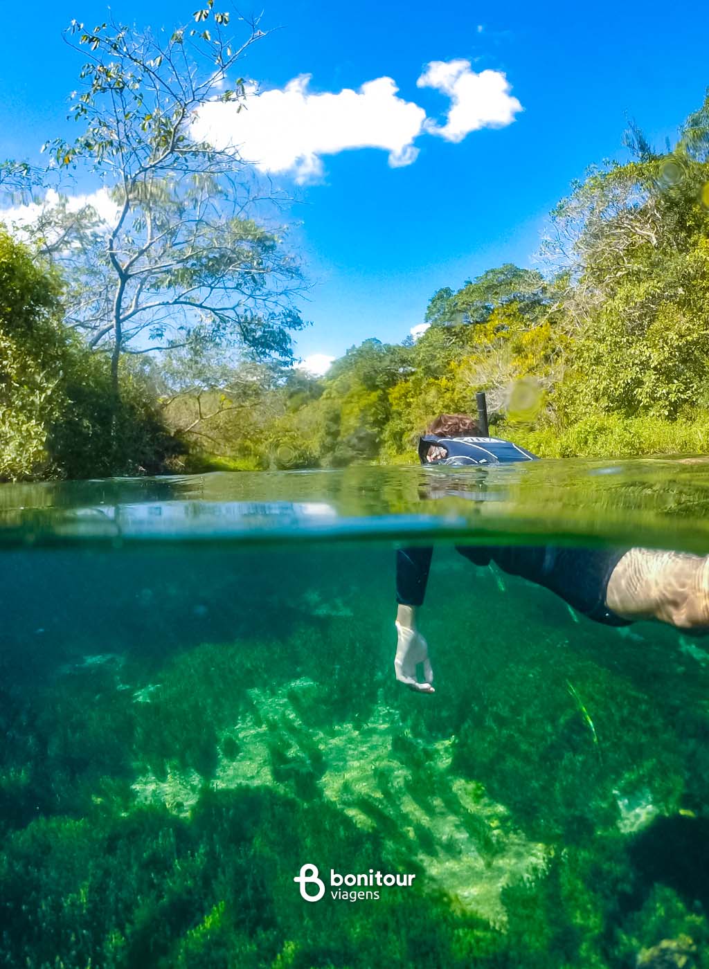Visão de dentro e fora da água, de pessoa praticando flutuação, com céu ensolarado no Rio Sucuri.