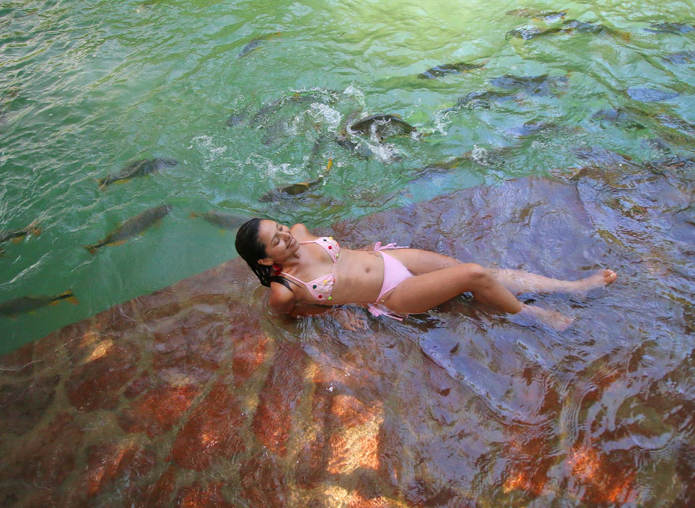 Mulher com traje de banho deitada em pedra sob a água, com cardume de peixes ao lado.