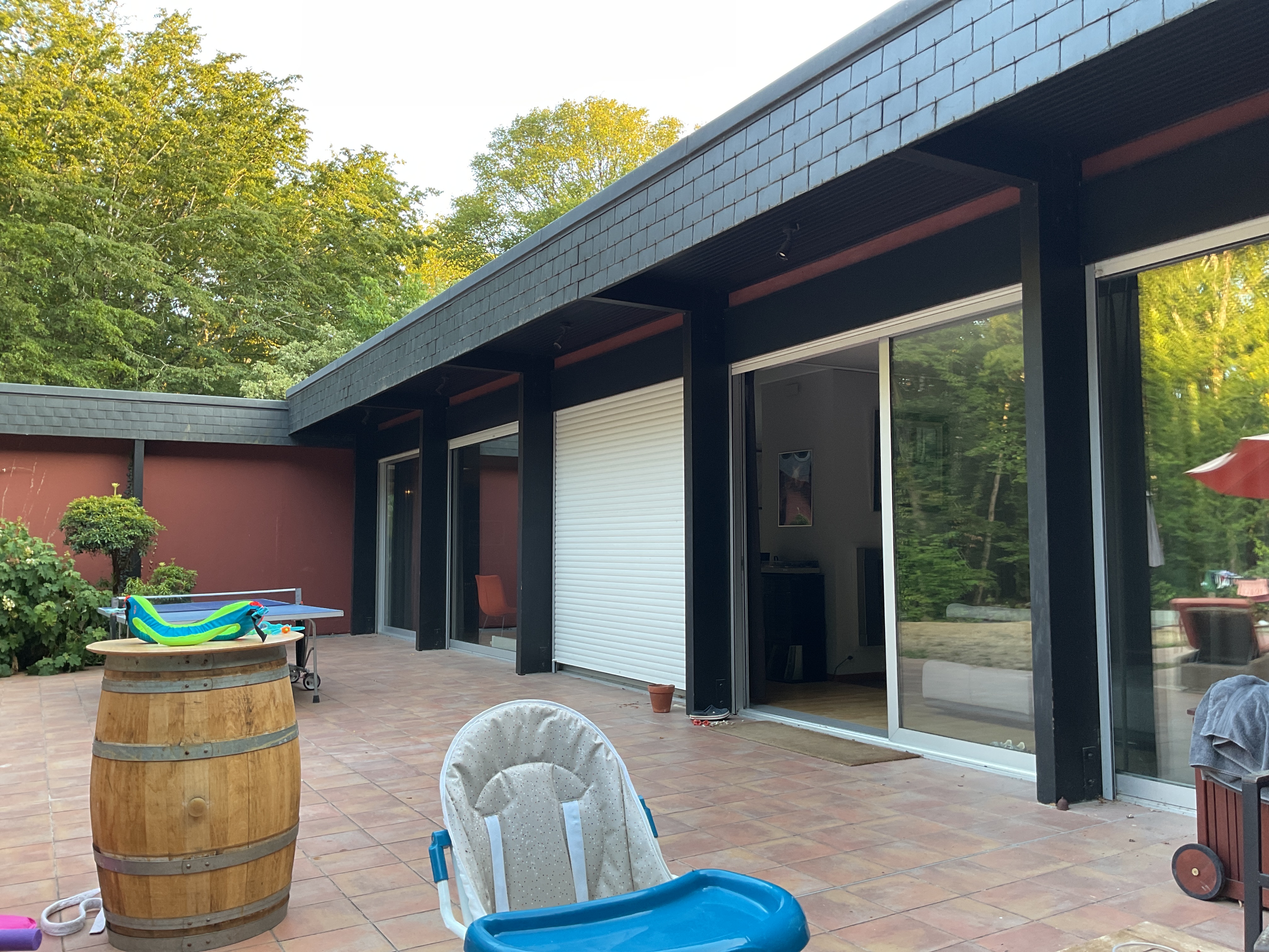 Exterior view of modern home with black brick facade, terracotta patio, large sliding glass doors, and wooden barrel planter with surrounding gardens.