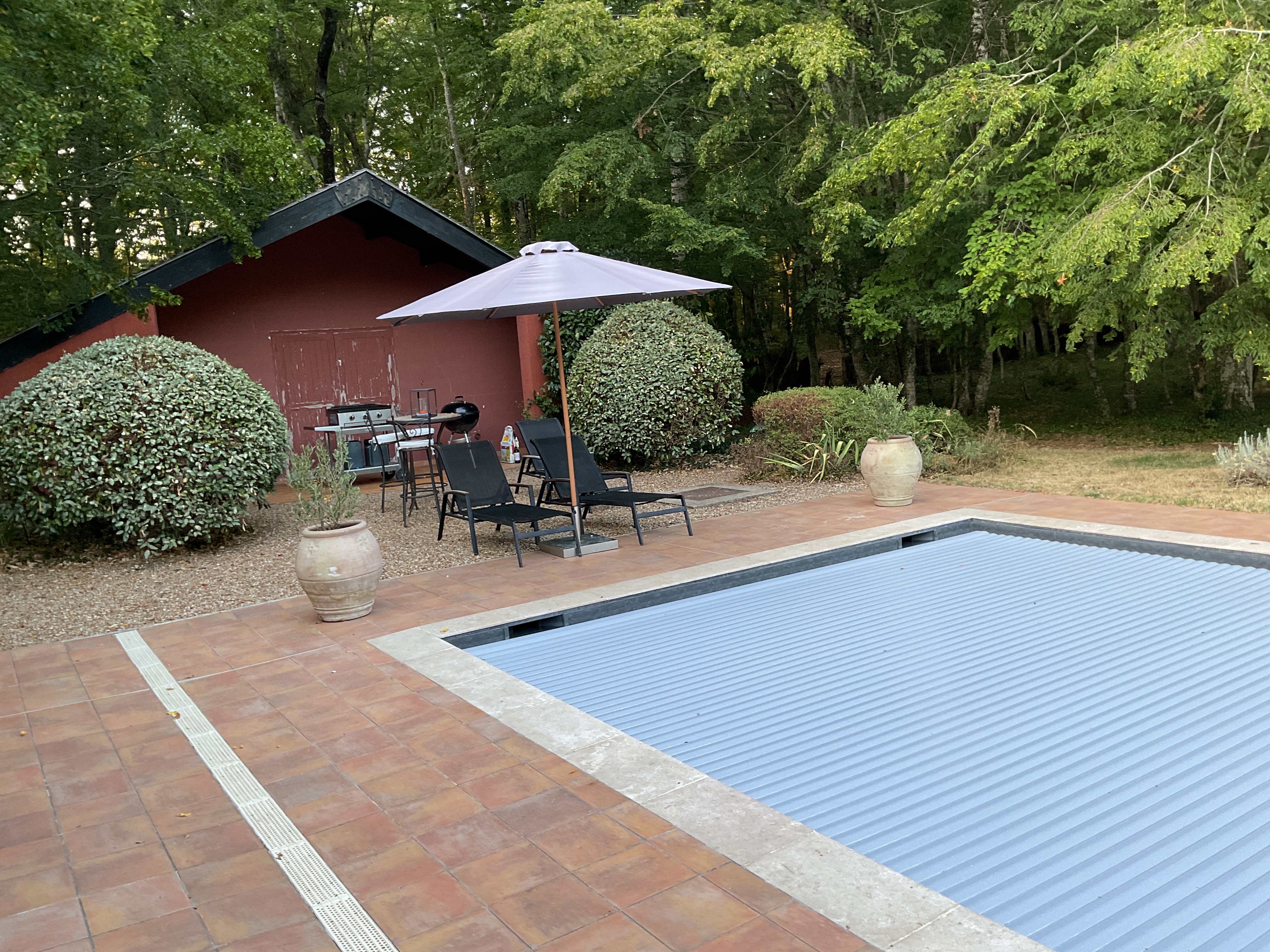 Exterior backyard view featuring a covered pool with blue rolling cover, terracotta patio, red brick pool house, lounge seating under umbrella, and lush wooded surroundings.