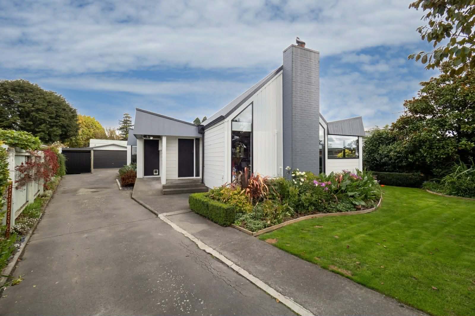 Exterior view of a modern white single-story house with gray chimney, large windows, concrete driveway, landscaped front garden, and attached garage.
