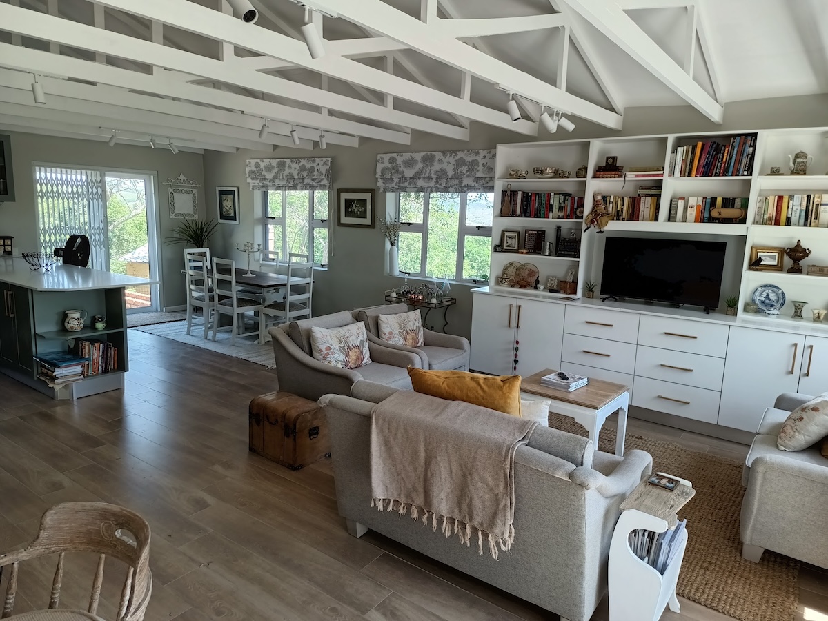 Interior living room with exposed white beam ceiling, built-in white shelving with TV, comfortable seating, dark hardwood floors, and abundant natural light from large windows.